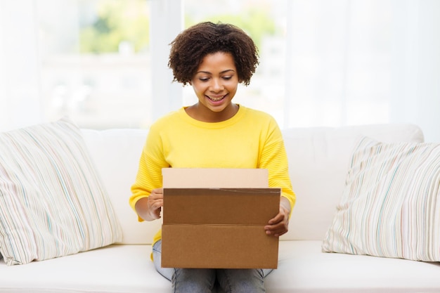 concept de personnes, de livraison, d'expédition et de service postal - jeune femme afro-américaine heureuse ouvrant une boîte en carton ou un colis à la maison