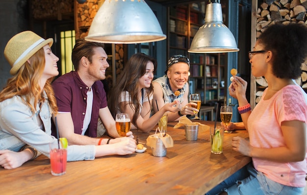 concept de personnes, de loisirs, d'amitié et de communication - groupe d'amis souriants heureux buvant de la bière et des cocktails mangeant et parlant au bar ou au pub