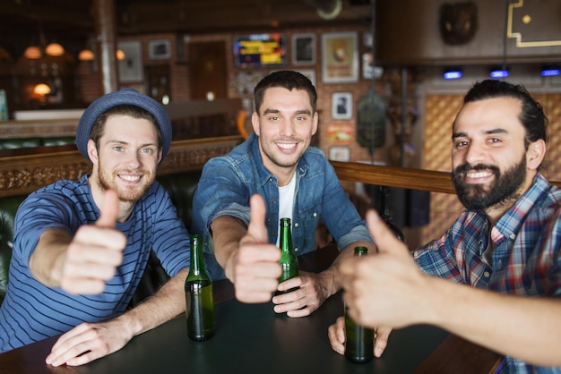 concept de personnes, de loisirs, d'amitié, de gestes et d'enterrement de vie de garçon - amis masculins heureux buvant de la bière en bouteille et montrant les pouces vers le haut au bar ou au pub