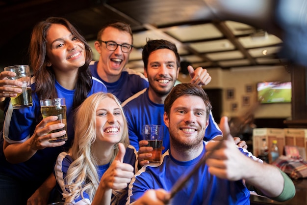 concept de personnes, de loisirs, d'amitié et de technologie - amis heureux ou fans de football prenant des photos avec un bâton de selfie de smartphone, buvant de la bière et montrant les pouces vers le haut au sport au bar ou au pub