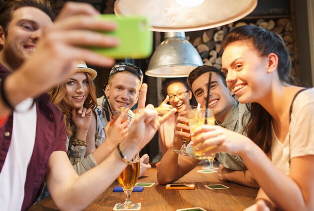 concept de personnes, de loisirs, d'amitié, de technologie et de communication - groupe d'amis souriants heureux avec un smartphone et des boissons prenant un selfie au bar ou au pub