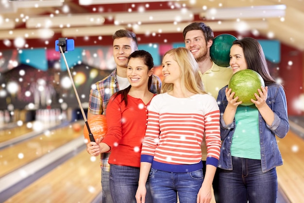 concept de personnes, de loisirs, de sport, d'amitié et de divertissement - amis heureux prenant un selfie avec un smartphone sur un monopode dans un club de bowling en hiver