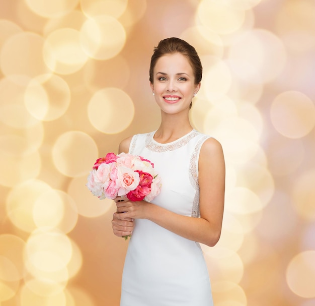 concept de personnes, de mariage, de vacances et de célébration - mariée souriante ou demoiselle d'honneur en robe blanche avec bouquet de fleurs sur fond de lumières beiges