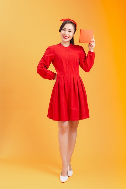 Concept de personnes, noël, anniversaire et vacances - heureuse jeune femme en robe rouge montrant/tenant avec boîte-cadeau sur fond orange.