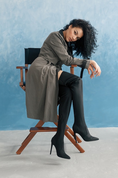 Concept de personnes, de race, d'ethnicité et de mode - modèle noir de beauté posant dans des bottes hautes. Jeune jolie belle femme mulâtre assis dans le fauteuil du réalisateur au film. femme en imperméable gris, coiffure afro