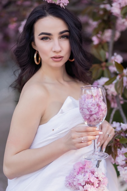 Concept de personnes, de repos, de confort et de loisirs - Jeune femme dans les rues du soir enveloppée dans une couverture. Belle jeune fille dans le parc et un sakura d'arbre fleuri tenant un verre avec des pétales