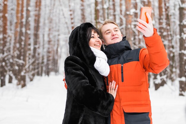 Concept de personnes, de saison, d'amour, de technologie et de loisirs - couple heureux prenant une photo avec un bâton de selfie pour smartphone sur fond d'hiver