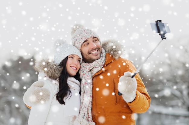 Concept de personnes, de saison, d'amour, de technologie et de loisirs - couple heureux prenant une photo avec un bâton de selfie pour smartphone sur fond d'hiver