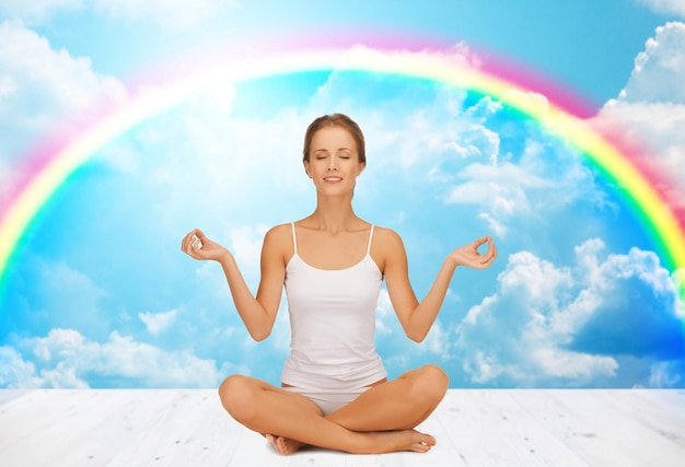 concept de personnes, de santé, de bien-être et de méditation - femme en sous-vêtements méditant dans la pose de lotus de yoga sur un plancher en bois sur des nuages blancs et un arc-en-ciel sur fond de ciel bleu