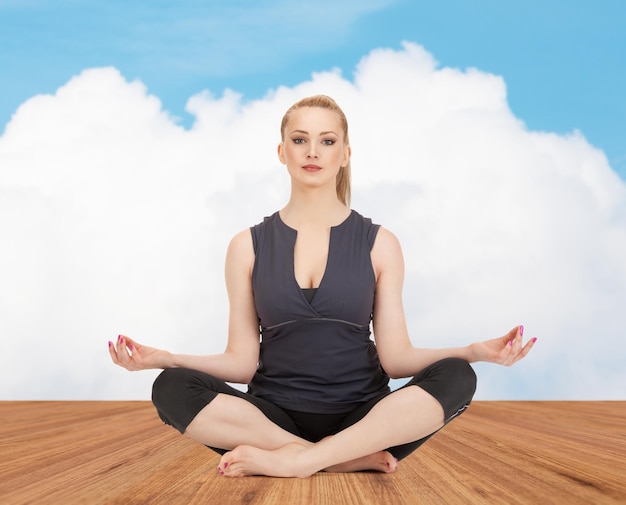 concept de personnes, de santé, de bien-être et de méditation - heureuse jeune femme méditant dans la pose de lotus de yoga sur un sol en bois sur fond de nuages blancs et de ciel bleu