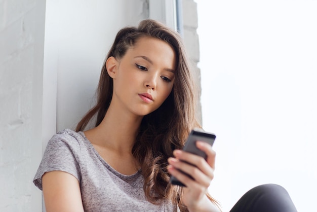 concept de personnes, de technologie et d'adolescents - triste malheureuse jolie adolescente assise sur le rebord de la fenêtre avec un smartphone et des SMS