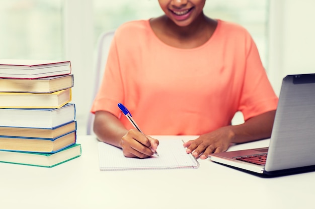 concept de personnes, de technologie, d'apprentissage et d'éducation - gros plan d'une jeune femme afro-américaine heureuse assise à table avec un ordinateur portable et des livres et écrivant sur un ordinateur portable à la maison