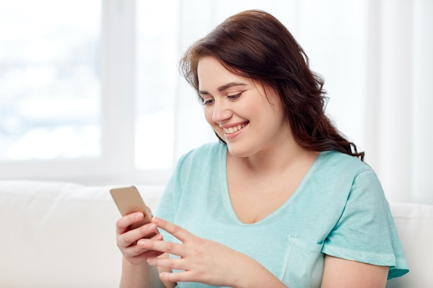 concept de personnes, de technologie, de communication et de loisirs - heureuse jeune femme de taille plus assise sur un canapé avec un smartphone à la maison