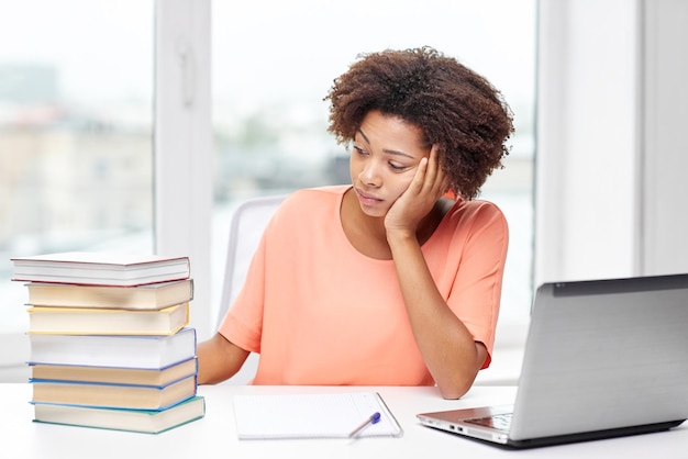 concept de personnes, de technologie et d'éducation - jeune femme afro-américaine ennuyée assise à table avec un ordinateur portable, des livres et un bloc-notes à la maison