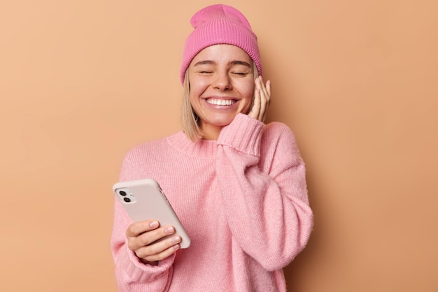 Concept de personnes et de technologie. Une femme positive sourit largement garde les yeux fermés attend un appel important a une expression heureuse touche le visage porte doucement un pull doux isolé sur fond marron