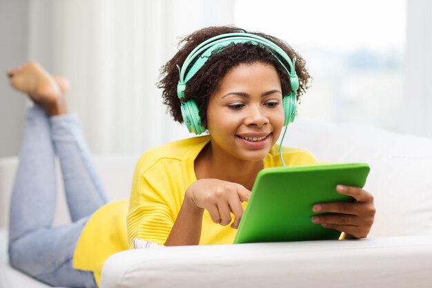 concept de personnes, de technologie et de loisirs - heureuse jeune femme afro-américaine allongée sur un canapé avec un ordinateur tablette et des écouteurs écoutant de la musique à la maison