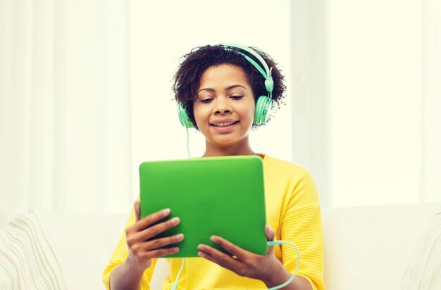 concept de personnes, de technologie et de loisirs - jeune femme afro-américaine heureuse assise sur un canapé avec un ordinateur tablette et des écouteurs écoutant de la musique à la maison