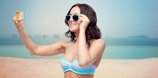 concept de personnes, de technologie, de voyage, de tourisme et d'été - heureuse jeune femme en maillot de bain bikini et lunettes de soleil prenant selfie avec smatphone sur la piscine à débordement sur fond de bord de mer