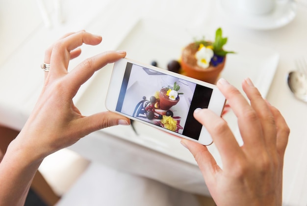 concept de personnes, de vacances, de technologie, de nourriture et de style de vie - gros plan d'une femme avec un smartphone prenant une photo de dessert au restaurant