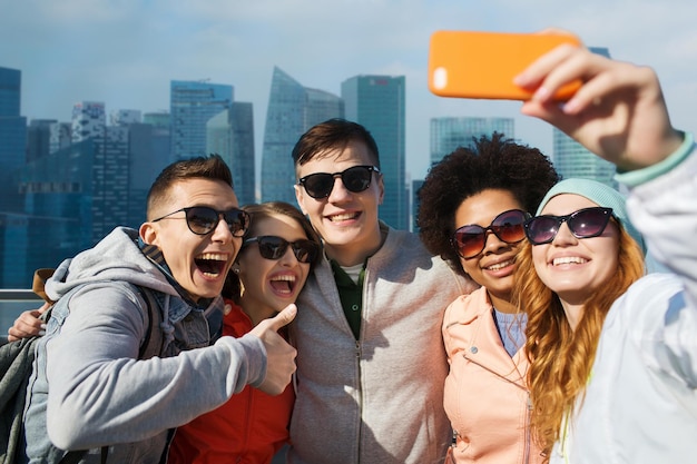 concept de personnes, de voyage, de tourisme, d'amitié et de technologie - groupe d'amis adolescents heureux prenant un selfie avec un smartphone et montrant les pouces vers le haut sur le fond de la ville de singapour