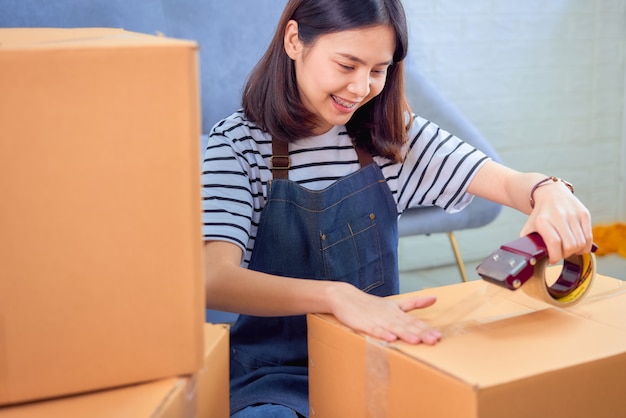 Concept de petite entreprise de démarrage, jeune femme asiatique propriétaire travaillant et emballant sur la boîte au client au canapé dans le bureau à domicile, le vendeur prépare la livraison.