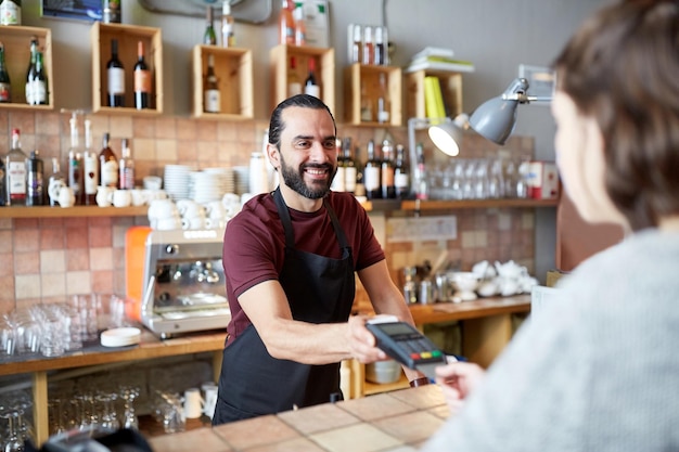 concept de petite entreprise, de personnes et de service - homme heureux ou serveur en tablier avec lecteur de carte et client payant au bar du café