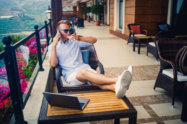 Concept de pigiste Jeune homme d'affaires prospère travaillant sur un ordinateur portable assis sur la terrasse en buvant du café et en parlant au téléphone