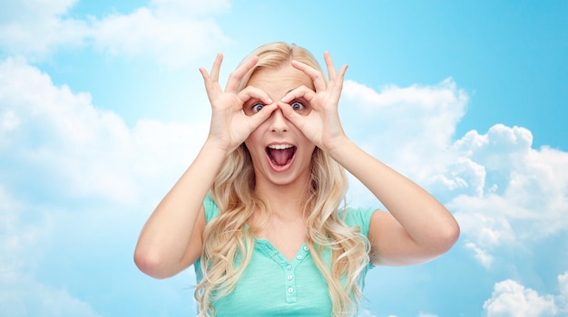 concept de plaisir, d'émotions, d'expressions et de personnes - jeune femme ou adolescente souriante regardant à travers des lunettes faites de doigts sur fond bleu ciel et nuages