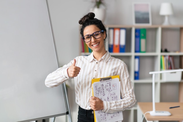 Concept de présentation happy female office worker près de tableau blanc vide vide montrant le pouce vers le haut et