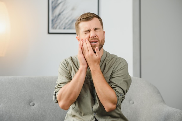 Concept de problèmes de santé dentaire Homme souffrant d'un fort mal de dents Patient masculin bouleversé avec un abcès nerveux enflammé ou une sensibilité des dents assis sur un canapé et touchant la joue avec une grimace de douleur