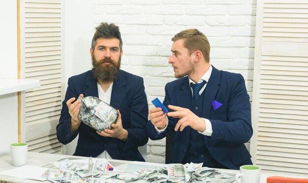 Concept de problèmes de trésorerie. Des collègues collectent de l'argent dans un bocal au lieu d'un compte bancaire. Partenaires commerciaux, hommes d'affaires lors d'une réunion au bureau. Manager avec barbe et collègue avec pot d'argent et carte