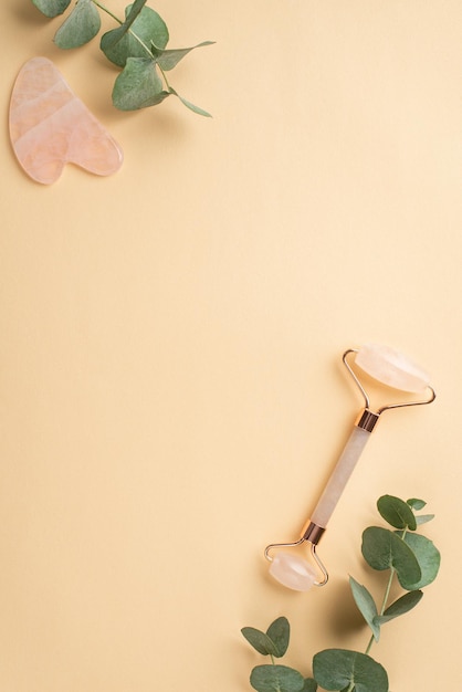 Concept de procédure de beauté Vue de dessus photo verticale du rouleau de quartz rose masseur gua sha et eucalyptus sur fond beige pastel isolé avec fond au milieu