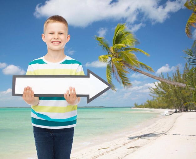 concept de publicité, de direction, d'été, de voyage et d'enfance - petit garçon souriant avec une flèche blanche vierge sur fond de plage tropicale