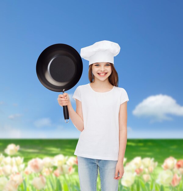 concept de publicité, d'enfance, de cuisine et de personnes - fille souriante en t-shirt blanc et chapeau de cuisine tenant la casserole sur fond de champ de fleurs