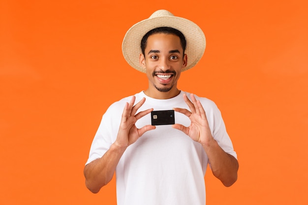 Concept de publicité, vacances et finances. Tout ce dont vous avez besoin, c'est de cette banque. Gai homme afro-américain souriant en chapeau d'été, t-shirt blanc, tenant la carte de crédit, mur orange debout