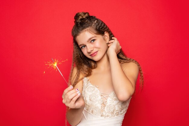 Concept de pyrotechnie et de personnes - jeune femme souriante ou femme heureuse adolescente avec des cierges magiques célèbrent en robe blanche sur fond rouge.