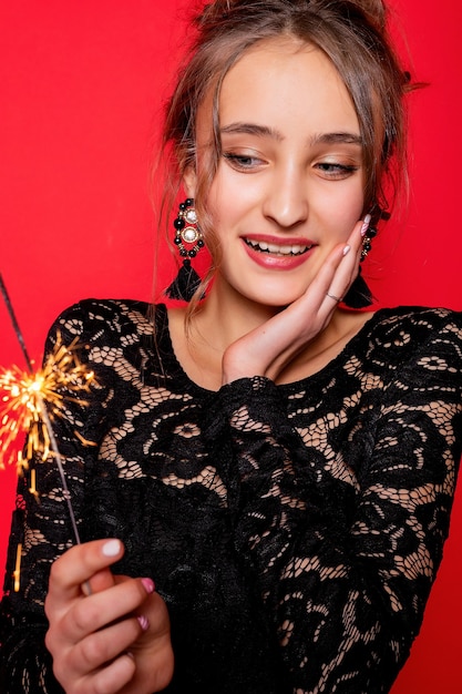 Photo concept de pyrotechnie et de personnes - jeune femme souriante ou femme heureuse adolescente avec des cierges magiques célèbrent en robe noire sur fond rouge. la fille est surprise