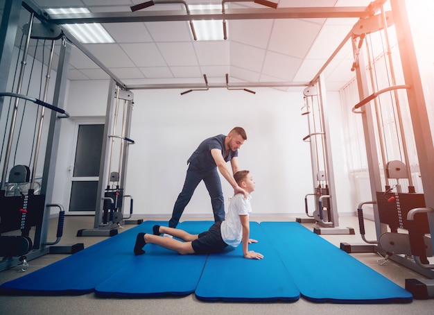 Concept de réadaptation. Jeune garçon faisant des exercices sur tapis sous la supervision d'un physiothérapeute