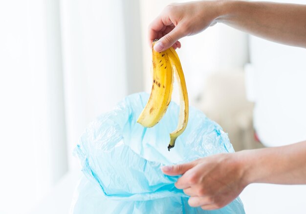 concept de recyclage, de déchets alimentaires, d'ordures, d'environnement et d'écologie - gros plan de la main mettant la peau de banane dans un sac poubelle à la maison