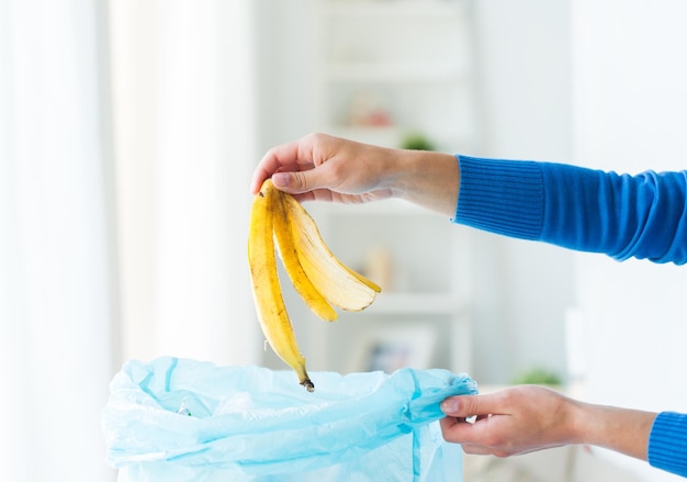 concept de recyclage, de déchets alimentaires, d'ordures, d'environnement et d'écologie - gros plan de la main mettant la peau de banane dans un sac poubelle à la maison