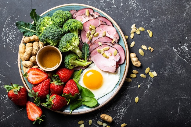 Concept de régime cétogène à faible teneur en glucides, vue de dessus. Assiette sur fond noir en pierre avec aliments céto : œuf, viande, huile d'olive, brocoli, baies, noix, graines, épinards. Graisses saines, alimentation saine pour perdre du poids