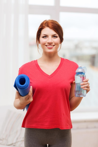 concept de remise en forme, de maison et de régime - adolescente souriante avec une bouteille d'eau et un tapis de yoga après avoir fait de l'exercice à la maison