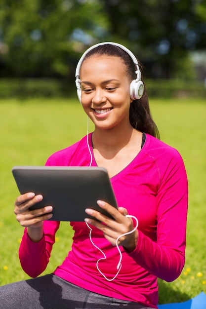 concept de remise en forme, de parc, de technologie et de sport - femme afro-américaine souriante avec ordinateur tablette et casque sur tapis à l'extérieur