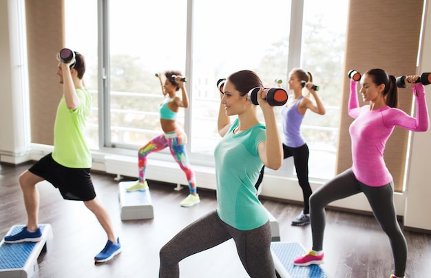 concept de remise en forme, de sport, d'aérobic et de personnes - groupe de personnes souriantes travaillant avec des haltères fléchissant les muscles sur des plates-formes de marche dans la salle de sport