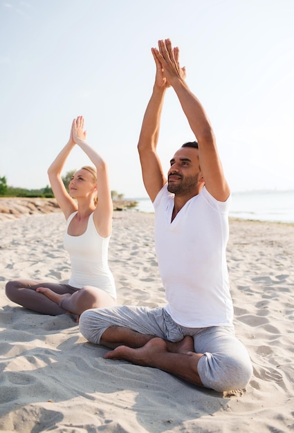 concept de remise en forme, de sport, d'amitié et de style de vie - couple souriant faisant des exercices de yoga assis sur le sable à l'extérieur