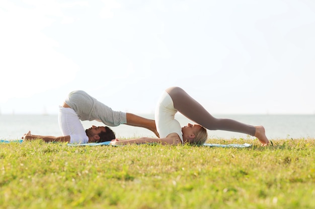 concept de remise en forme, de sport, d'amitié et de style de vie - couple souriant faisant des exercices de yoga sur des tapis à l'extérieur