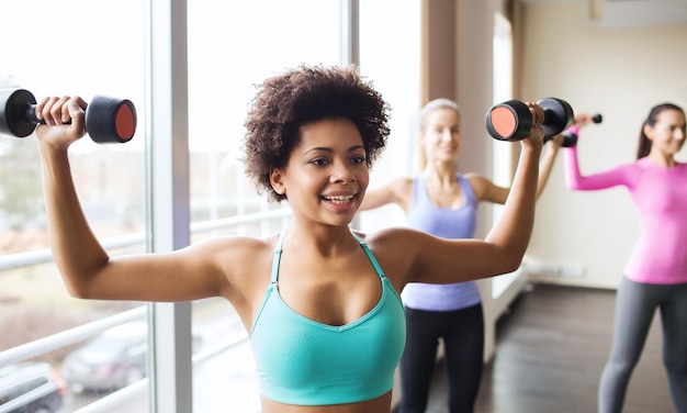 Concept de remise en forme, sport, formation et style de vie - groupe de femmes heureuses avec des haltères flexion des muscles dans la salle de gym