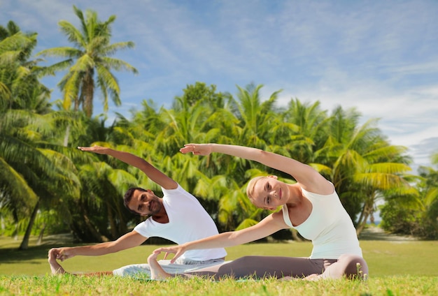 concept de remise en forme, de sport et de personnes - couple faisant des exercices de yoga en plein air sur fond naturel exotique avec des palmiers