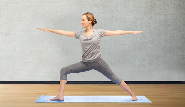 concept de remise en forme, de sport, de personnes et de mode de vie sain - femme faisant une pose de guerrier de yoga sur un tapis sur fond de salle ou de salle de sport