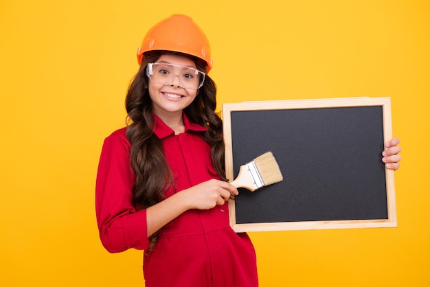 Concept de rénovation et de construction pour enfants Petite fille dans un casque de protection et un pinceau pour peindre des murs isolés sur fond jaune Visage heureux émotions positives et souriantes d'une adolescente